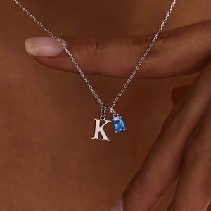 以下が含まれることがあります： 繊細なチェーンが付いたシルバーのネックレス。ネックレスには、小さな大文字の「K」ペンダントと、長方形のライトブルーの宝石ペンダントが付いています。ネックレスは着用されています。