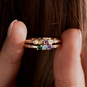 May include: Two gold rings, each with a row of colourful gemstones. The top ring features a yellow, green, and white stone, while the bottom ring has green, purple, and white stones. The rings are held up by a person's fingers.
