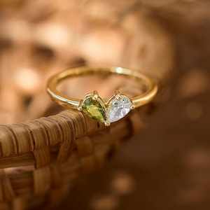 May include: A gold ring with two pear-shaped gemstones, a green and a white one. The ring is set against a blurred background. The ring is placed on a woven basket. The ring's design and craftsmanship are highlighted.