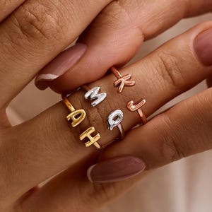 May include: Close-up of several rings with individual letter designs. The rings are in gold, silver, and rose gold tones. The letters visible are A, H, M, Q, K, and J. The rings are worn on a finger.