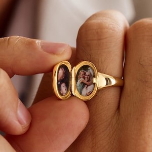 May include: A gold-colored locket ring with an oval-shaped compartment that opens to reveal two small photographs. The ring is held open, displaying the images inside. The ring is on a finger.
