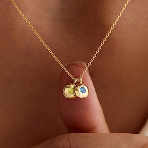 Könnte beinhalten: Eine goldene Halskette mit zwei kleinen, runden Goldanhängern. Jeder Anhänger ist mit einem einzelnen Edelstein besetzt, einer in leuchtendem Limettengrün und der andere in hellem Blau. Die Kette ist zart und hängt an einer dünnen Goldkette.