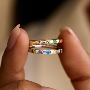 Puede incluir: Dos anillos apilados con pequeñas piedras preciosas redondas y multicolores. El anillo superior es de tono dorado con tres piedras, mientras que el inferior es de tono plateado con cinco piedras de colores azul, amarillo, morado y verde.
