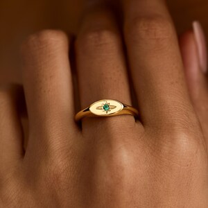 Könnte beinhalten: Ein goldener Siegelring mit einer ovalen Ringplatte und polierter Oberfläche. In der Mitte des Rings ist ein Sternmotiv eingraviert, das einen kleinen, runden, leuchtend grünen Edelstein enthält, der direkt in die Metalloberfläche eingelassen ist.