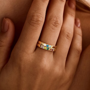 May include: Two stacked gold rings featuring a series of small, multi-colored gemstones set into the bands. The stones include shades of blue, green, yellow, and red, creating a vibrant, linear pattern across the surface of each polished gold ring.