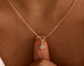 May include: A gold-toned necklace featuring a delicate chain and a small, star-shaped pendant. The center of the star is set with a single, round, bright blue gemstone. The pendant hangs from a thin, polished gold-colored metal chain.