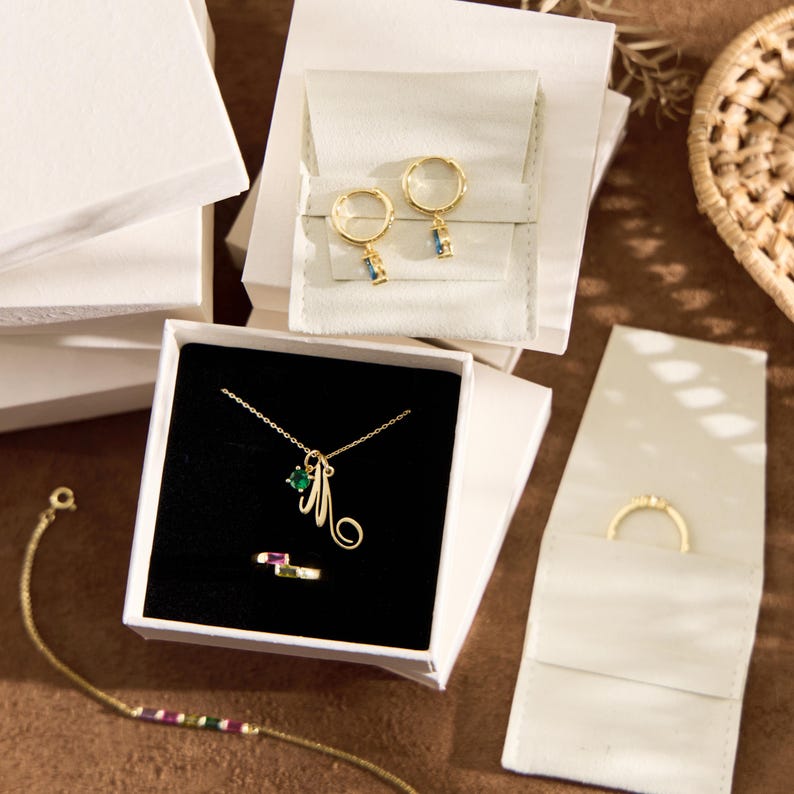 May include: An assortment of gold-toned jewelry, including a necklace with a green gem and initial pendant, hoop earrings with blue and green accents, and a ring with a rectangular gem. The jewelry is displayed in white boxes and pouches.