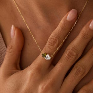 May include: A delicate gold chain necklace featuring two small heart-shaped stones set side by side. One stone is a deep olive green, and the other is a clear, sparkling white. The pendant is suspended from a thin, polished gold-tone metal chain.