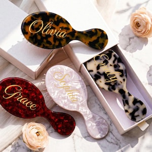 Puede incluir: Cuatro cepillos para el cabello personalizados en varios colores y patrones, cada uno con un nombre en escritura dorada. Los cepillos tienen diseños de carey, rojo, blanco y blanco y negro. Los cepillos están en cajas blancas.