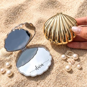 Pode incluir: Um espelho de bolso dourado em forma de concha com design articulado. Um lado apresenta uma superfície refletora com o nome Olivia impresso em letra cursiva preta. O outro lado é uma concha dourada rígida e estriada. Várias pérolas brancas estão espalhadas.