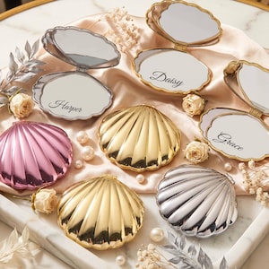 Pode incluir: Uma coleção de espelhos de bolso em forma de concha com acabamentos metálicos dourados, prateados e rosa. Alguns espelhos estão abertos, revelando superfícies refletoras com os nomes Harper, Daisy e Grace impressos em letra cursiva preta.