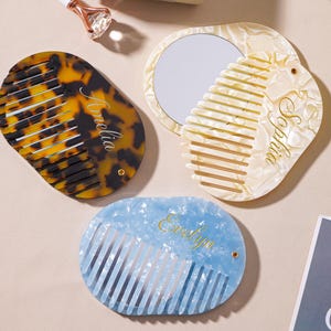 May include: Three personalised combs are displayed. One is tortoiseshell with "Amelia" in gold script, another is pearlescent with a mirror and "Sophia", and the third is blue with "Evelyn".