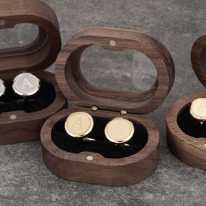 May include: Three wooden boxes with lids, each containing a pair of cufflinks. The cufflinks are round and have a single letter engraved on them. The boxes are made of dark wood and have a polished finish.