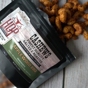 May include: A black resealable pouch of High Desert Provisions Applewood BBQ cashews with a net weight of 8 oz. The label features the HDP logo, text describing the salted and roasted nuts, and a pile of seasoned brown cashews spilling out.
