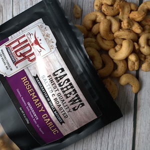 May include: A black pouch of High Desert Provisions Rosemary Garlic cashews with a clear window showing the nuts. The label reads Cashews, Finest Quality, Salted & Roasted, Handcrafted in Small Batches, Net Wt 8 oz (226g). Loose cashews are nearby.