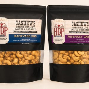 May include: Two black stand-up pouches of High Desert Provisions cashews. The left bag is labeled Backyard BBQ with a blue accent, and the right bag is labeled Rosemary Garlic with a purple accent. Each 8 oz (226g) bag features a clear window.