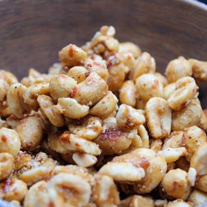 May include: A wooden bowl filled with roasted peanuts, coated in a sweet and savory glaze. The peanuts are a light brown color and have a slightly crunchy texture.