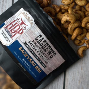 May include: A black plastic pouch of High Desert Provisions Backyard BBQ cashews. The label features the text CASHEWS, FINEST QUALITY, SALTED & ROASTED, BACKYARD BBQ, and lists ingredients. Some seasoned roasted cashews are spilled outside the bag.