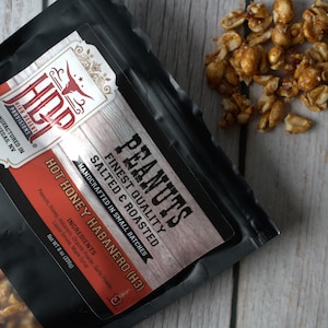 May include: A black plastic pouch of High Desert Provisions Hot Honey Habanero peanuts. The label features a bull skull logo, text reading Peanuts Finest Quality Salted & Roasted, and a net weight of 8 oz (226g). Loose roasted peanuts are visible.