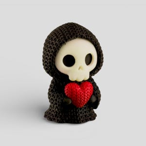 A small, knitted figurine of a skull wearing a black hooded cloak and holding a red heart.