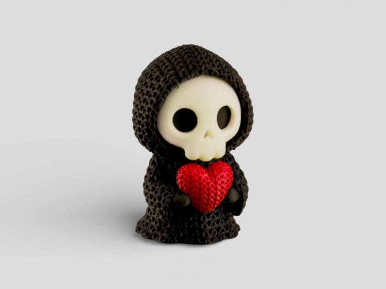 A small, knitted figurine of a skull wearing a black hooded cloak and holding a red heart.