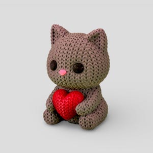 A small, brown, knitted stuffed animal cat holding a red heart.