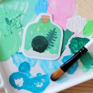 May include: A green glass bottle sticker with a black orb, green leaves, and white sparkles on a white ceramic paint palette with various colors of paint.