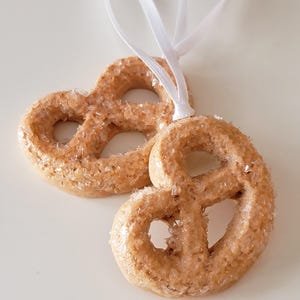 May include: Two pretzel-shaped ornaments with a light brown color and a textured, crystallized surface. Each ornament is suspended by a white ribbon. The ornaments are likely for holiday or seasonal decor.