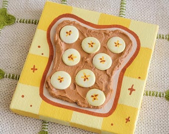 Puede incluir: Un lienzo cuadrado que muestra una rebanada de pan tostado con crema marrón y siete rodajas de plátano blancas con centros amarillos. El fondo es un patrón de cuadros amarillos y blancos con pequeños signos más naranjas.