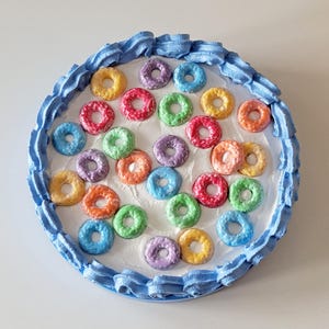 May include: A round, colorful cake with a blue frosting border and a white frosted center. The cake is decorated with various colored, ring-shaped candies, including yellow, pink, green, purple, orange, and blue. The cake is a whimsical and playful dessert.