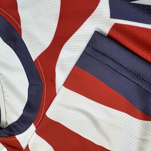 May include: Close-up of a red, white, and blue athletic jersey. The jersey features a pattern of red and white stripes with a blue collar and sleeve trim. The fabric has a mesh texture.