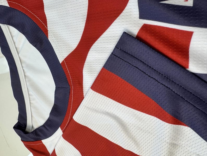 May include: Close-up of a red, white, and blue athletic jersey. The jersey features a pattern of red and white stripes with a blue collar and sleeve trim. The fabric has a mesh texture.