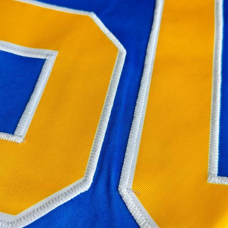 May include: Close-up of a blue fabric with the number "21" in yellow, outlined with white stitching. The number is large and prominent, taking up most of the frame. The fabric has a slight texture.