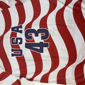 May include: A red, white, and blue USA jersey with the number 43. The jersey has wavy red stripes and the letters "USA" in navy blue. The collar is navy blue.