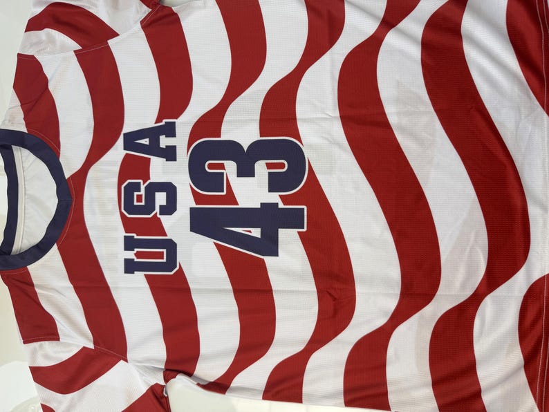 May include: A red, white, and blue USA jersey with the number 43. The jersey has wavy red stripes and the letters "USA" in navy blue. The collar is navy blue.