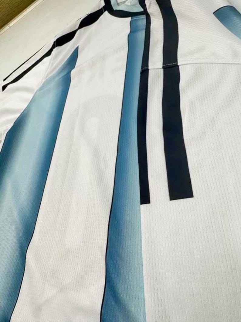 May include: White athletic jersey with blue and black vertical stripes. The jersey has a crew neck and a mesh-like texture. The stripes are a gradient of blue, fading from a darker shade to a lighter one.