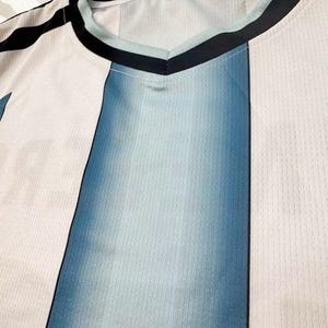 May include: Close-up of a V-neck jersey with a color block design. The jersey features vertical panels of white and blue, with a gradient effect in the blue sections. The neckline is trimmed with black and light blue accents.