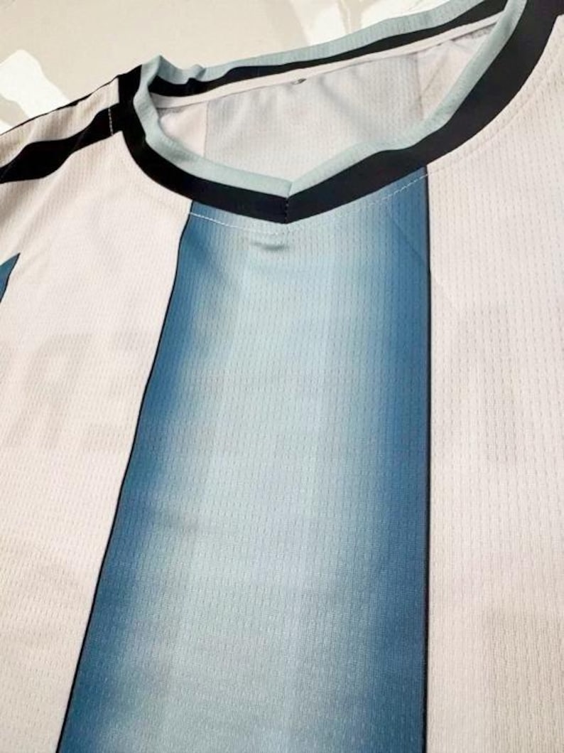 May include: Close-up of a V-neck jersey with a color block design. The jersey features vertical panels of white and blue, with a gradient effect in the blue sections. The neckline is trimmed with black and light blue accents.