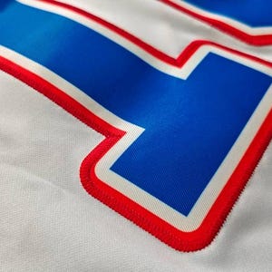 May include: Close-up of a white fabric with a large blue number outlined in red and white. The number has a textured appearance, with the red outline featuring a stitched design. The fabric has a subtle, woven texture.