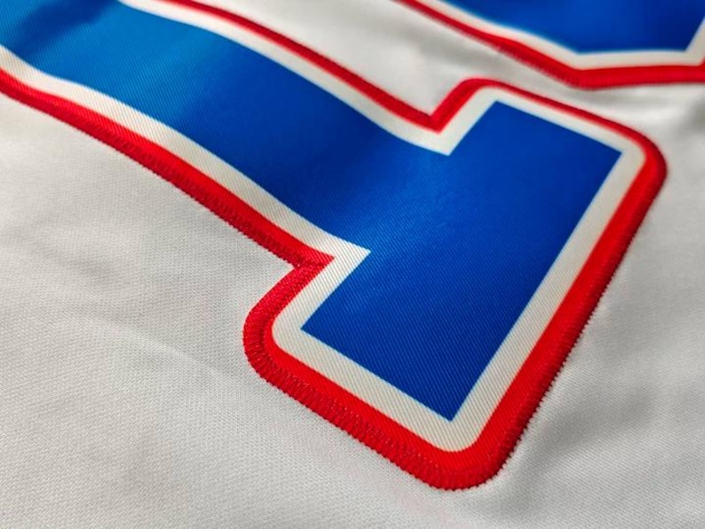 May include: Close-up of a white fabric with a large blue number outlined in red and white. The number has a textured appearance, with the red outline featuring a stitched design. The fabric has a subtle, woven texture.