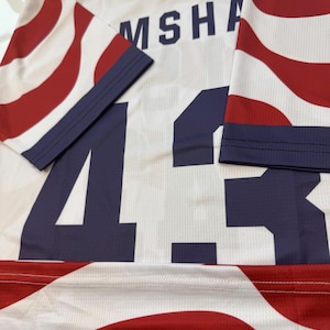 May include: White jersey with red and blue striped sleeves and accents. The number 43 is prominently displayed in navy blue, along with the name "MSHA" across the shoulders. The jersey has a mesh texture.