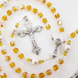 May include: A silver rosary with amber glass beads and pearl accents. The rosary features a silver crucifix and a silver chalice charm.