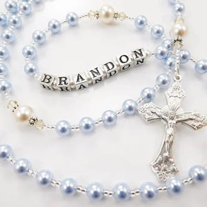 May include: A light blue rosary with pearl beads and a silver crucifix. The name "BRANDON" is spelled out in white lettered cubes. The rosary features a mix of light blue and cream-coloured beads.