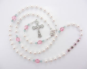 May include: A rosary featuring white pearl-like beads, translucent pink faceted beads, and small light blue accent beads. The piece includes a silver-toned metal crucifix, a central medal depicting a religious figure, and silver letter beads spelling JUNE.