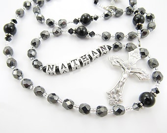 May include: A rosary featuring faceted dark gray beads, smooth black round beads, and silver-toned spacer beads. The rosary includes square silver-toned letter beads spelling the name NATHAN and a detailed silver-toned crucifix pendant.