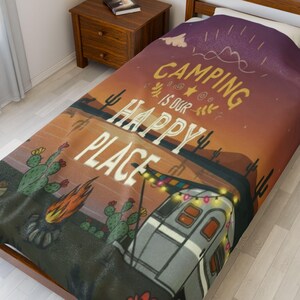 May include: A camping-themed blanket with a desert landscape design. The blanket features a camper, cacti, and a campfire, with the text "Camping is our Happy Place" in a stylized font. The color palette includes orange, purple, and green.