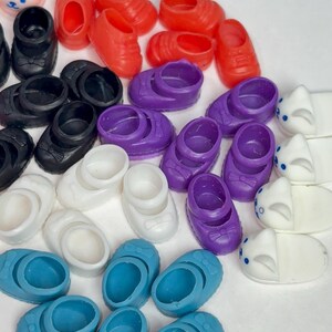 May include: A collection of miniature doll shoes in various colors, including red, black, white, purple, and blue. Some shoes have bows, while others are simple slip-ons. There are also several pairs of pink bunny slippers.