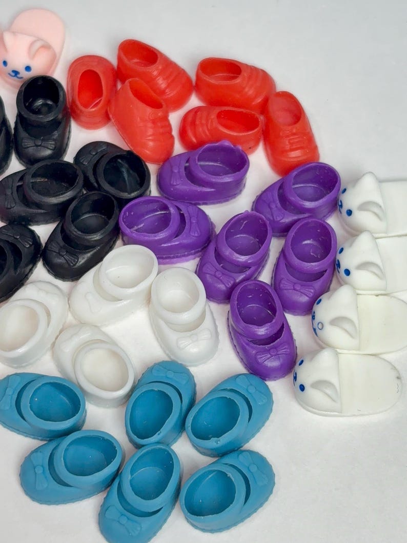 May include: A collection of miniature doll shoes in various colors, including red, black, white, purple, and blue. Some shoes have bows, while others are simple slip-ons. There are also several pairs of pink bunny slippers.