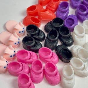May include: A collection of colorful miniature shoes in various styles. The assortment includes red sneakers, purple clogs, black Mary Janes with bows, pink shoes with bows, white slip-ons, and pink bunny slippers. These small shoes are likely for dolls or other toys.