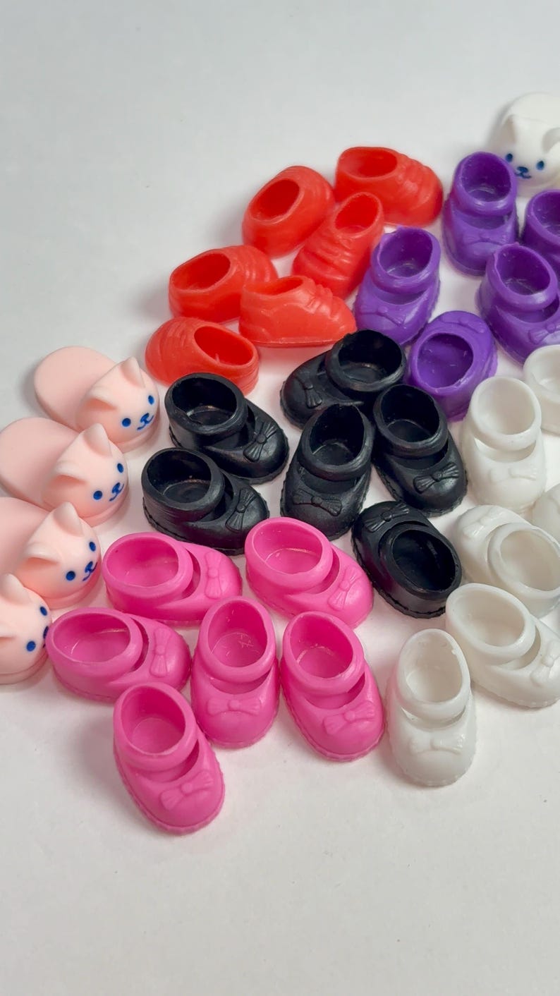 May include: A collection of colorful miniature shoes in various styles. The assortment includes red sneakers, purple clogs, black Mary Janes with bows, pink shoes with bows, white slip-ons, and pink bunny slippers. These small shoes are likely for dolls or other toys.